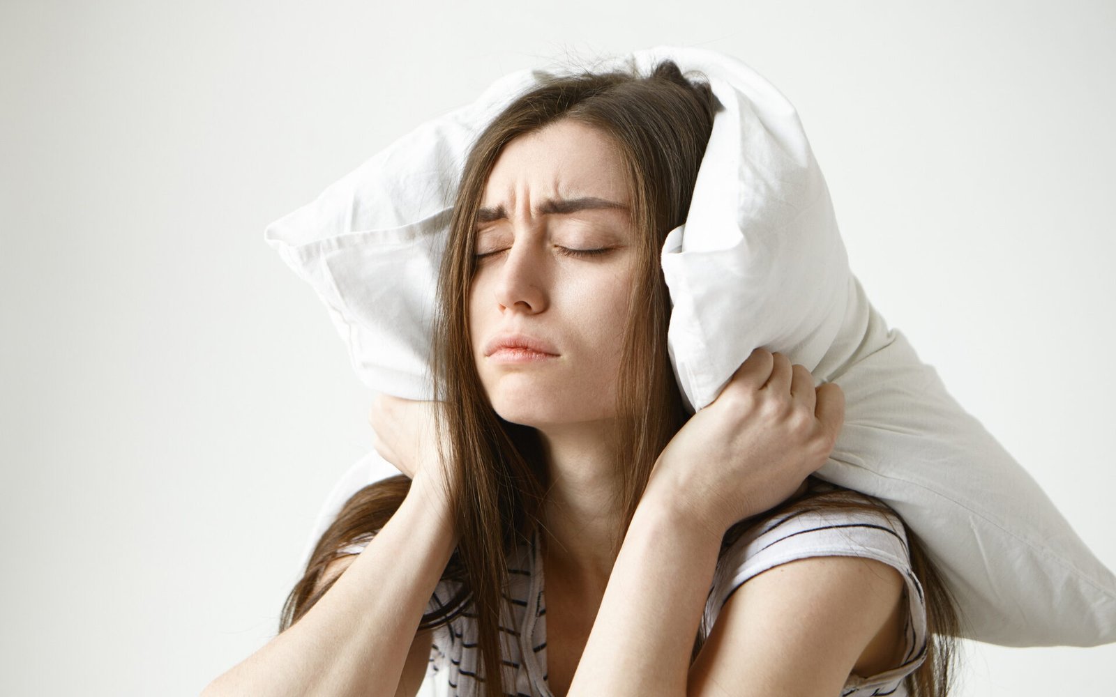 pretty-student-woman-in-striped-t-shirt-trying-to-have-some-sleep-sitting-in-bedroom-with-pillow-on-her-head-closing-eyes